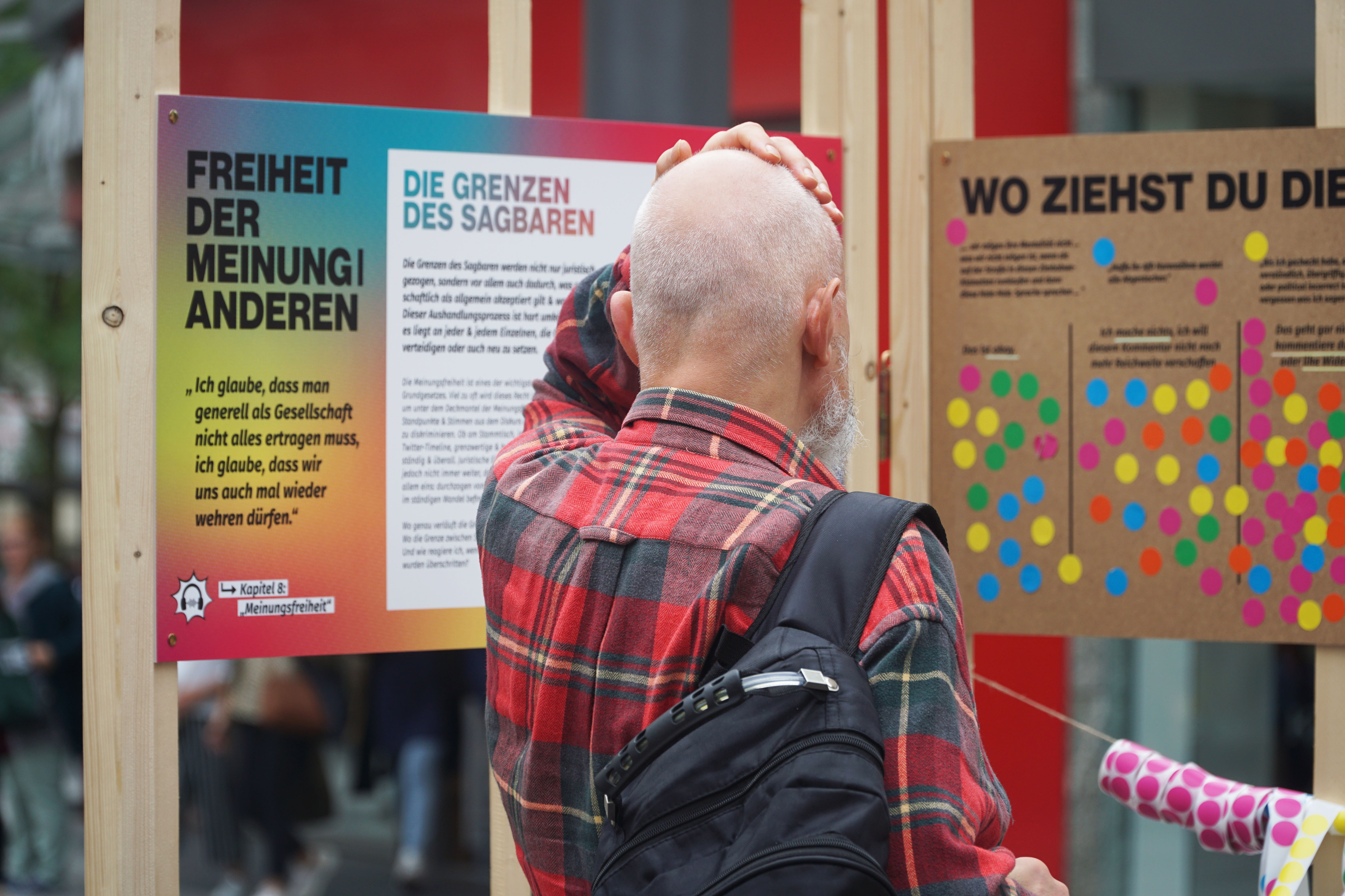 Ein Mann steht vor zwei Infotafel einer Ausstellung und fässt sich an den Kopf. Auf der linken Tafel steht groß "Freiheit der Meinung|Anderen" und die "Grenzen des Sagbaren". Auf der rechten Tafel steht "Wo ziehst du die Grenze", darunter sind fünf grenzwertige Aussagen aufgelistet und Möglichkeiten darauf zu reagieren.