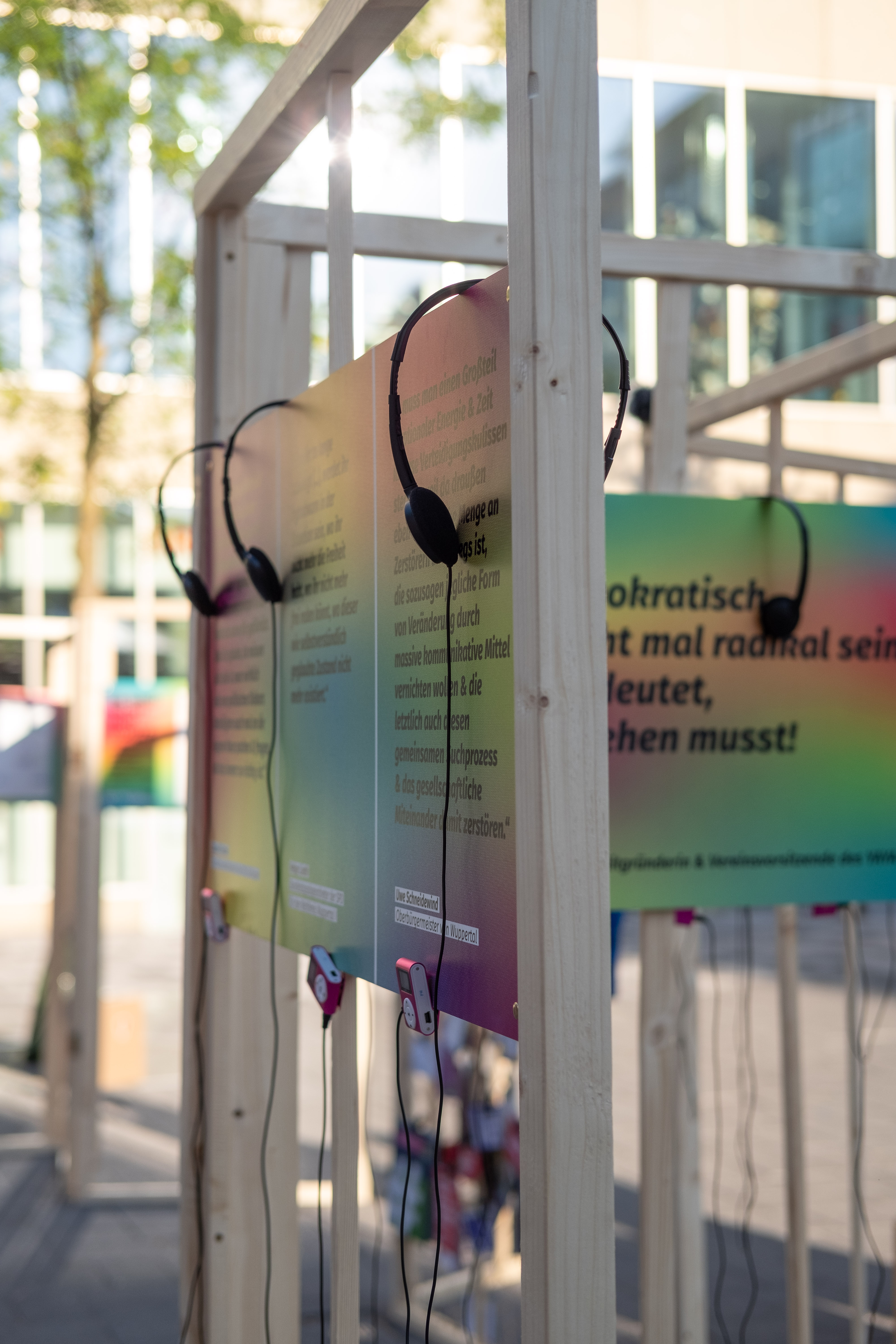 Es sind bunte Tafeln in Regenbogenfarben mit großen Zitaten abgebildet. An den Tafeln hängen MP3 Player und Kopfhörer.